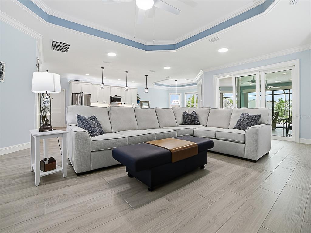 A living room features a large sectional sofa, a dark ottoman, and light wood flooring — 7004 EMILINE TERRACE, THE VILLAGES, FL