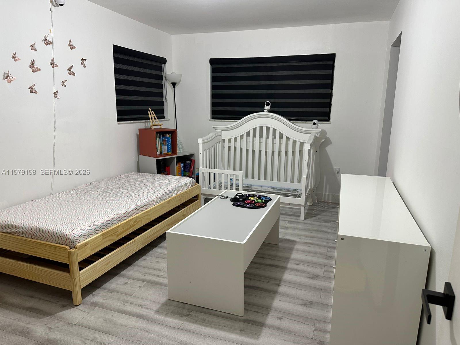 A bedroom features a wooden bed, a white crib, a central table, and light flooring — 931 NE 199th St # 22, Miami FL 33179, MIAMI, FL