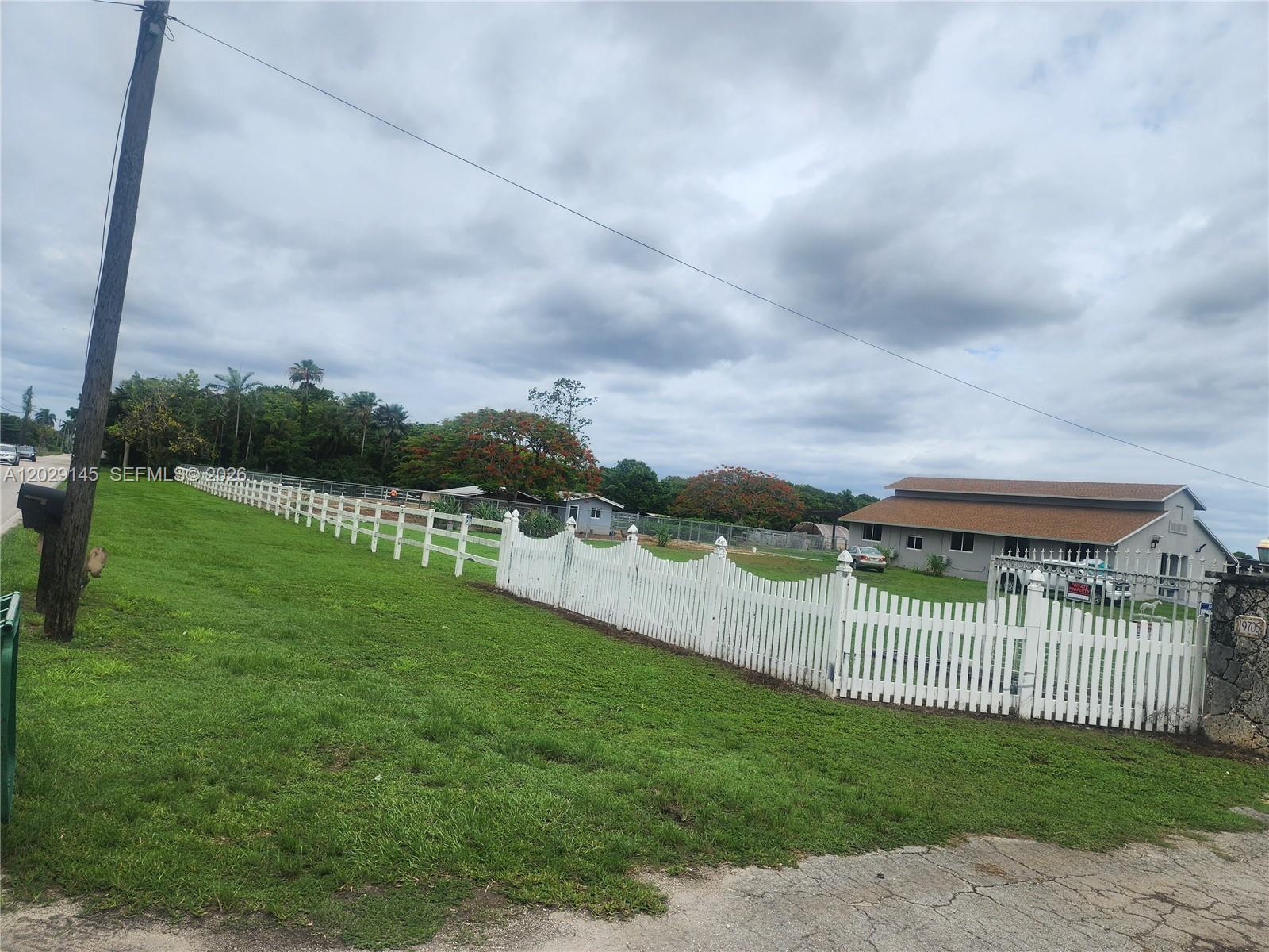 A white picket fence encloses a grassy yard in front of a house under clouds — HOMESTEAD, FL