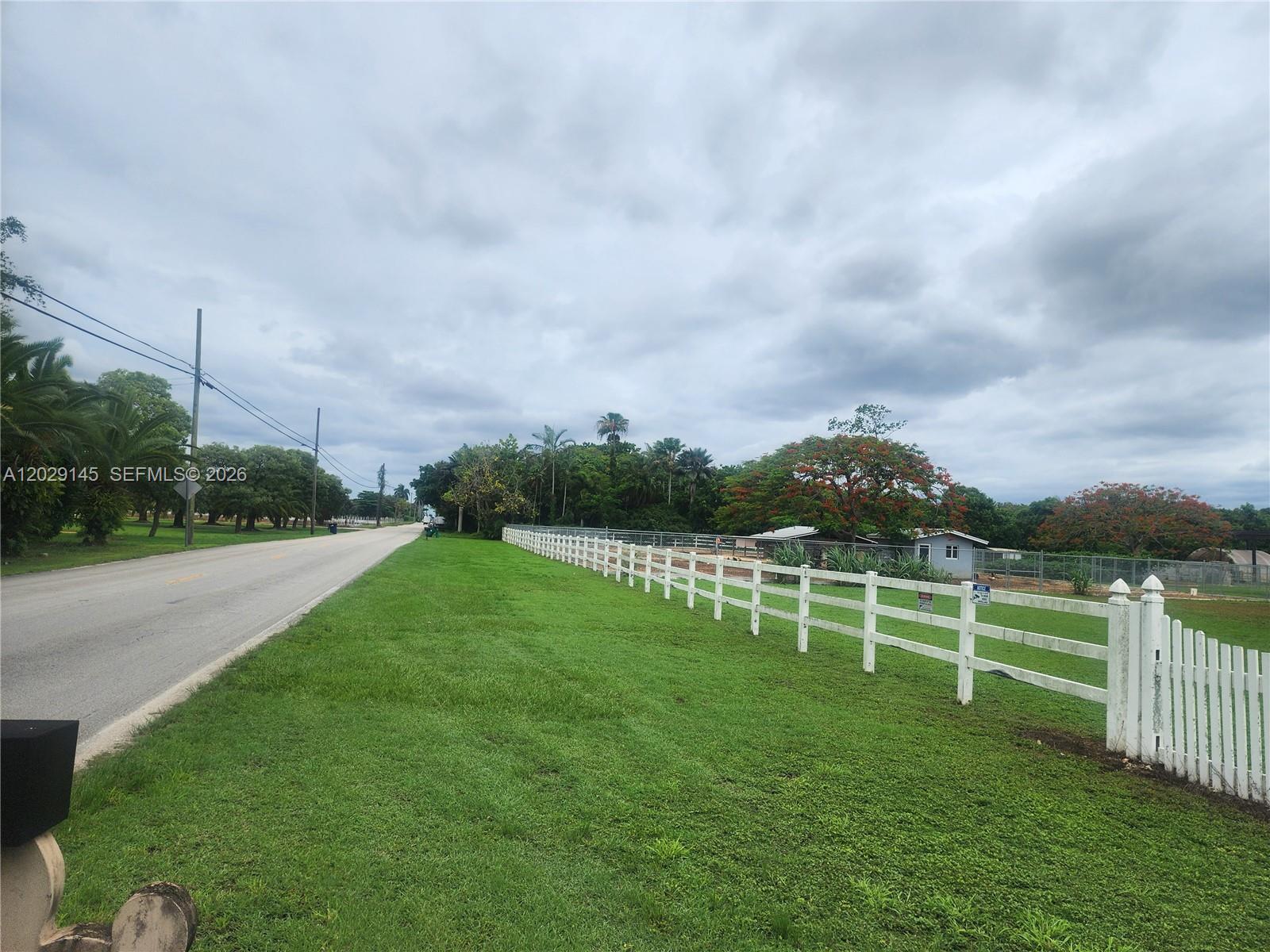 A white fence runs along a grassy field next to a paved road under cloudy skies — HOMESTEAD, FL