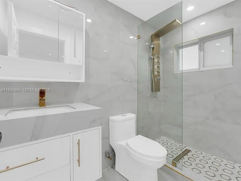 A modern bathroom features white marble-style tiles, a glass-enclosed shower, and gold-toned fixtures — 4900 NW 14th St, Lauderhill FL 33313, LAUDERHILL, FL