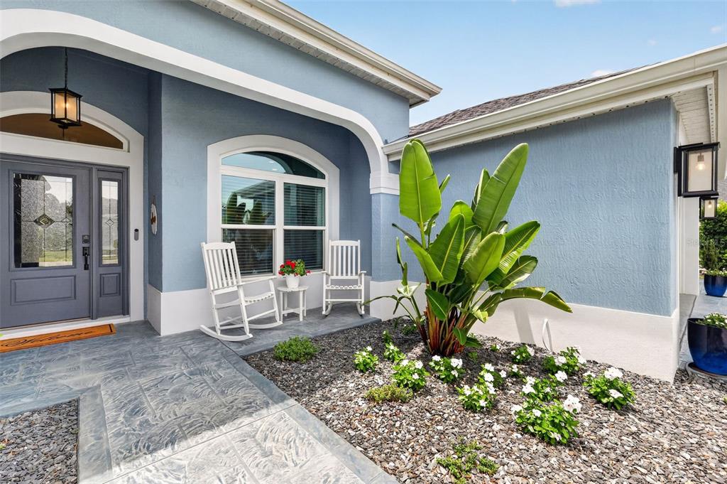 A blue house exterior features a covered porch with two white rocking chairs and landscaping — 6328 MERHOTTEIN LOOP, THE VILLAGES, FL