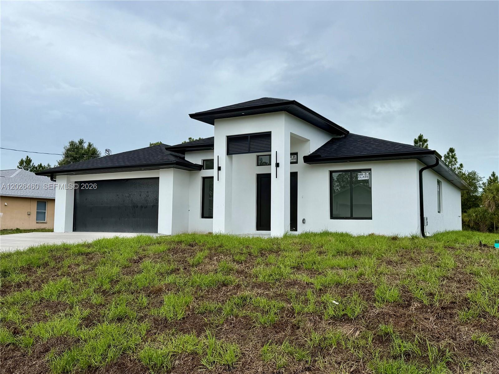 A modern, single-story white house with a black roof and garage door sits on grass — 911 Cleveland Ave, Lehigh Acres FL 33972, LEHIGH ACRES, FL