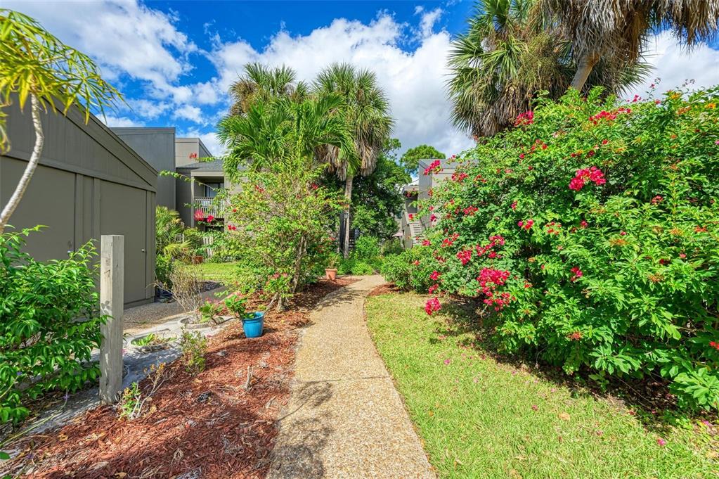 A concrete walkway leads between a building and a garden with flowering bushes and palms — 110 WOODLAND PLACE UNIT 110, OSPREY, FL