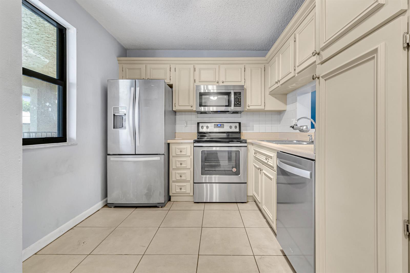 A kitchen with stainless steel appliances, off-white cabinets, light tile flooring, and a window — 5510 Lakeside Drive # 202, Margate FL 33063, MARGATE, FL