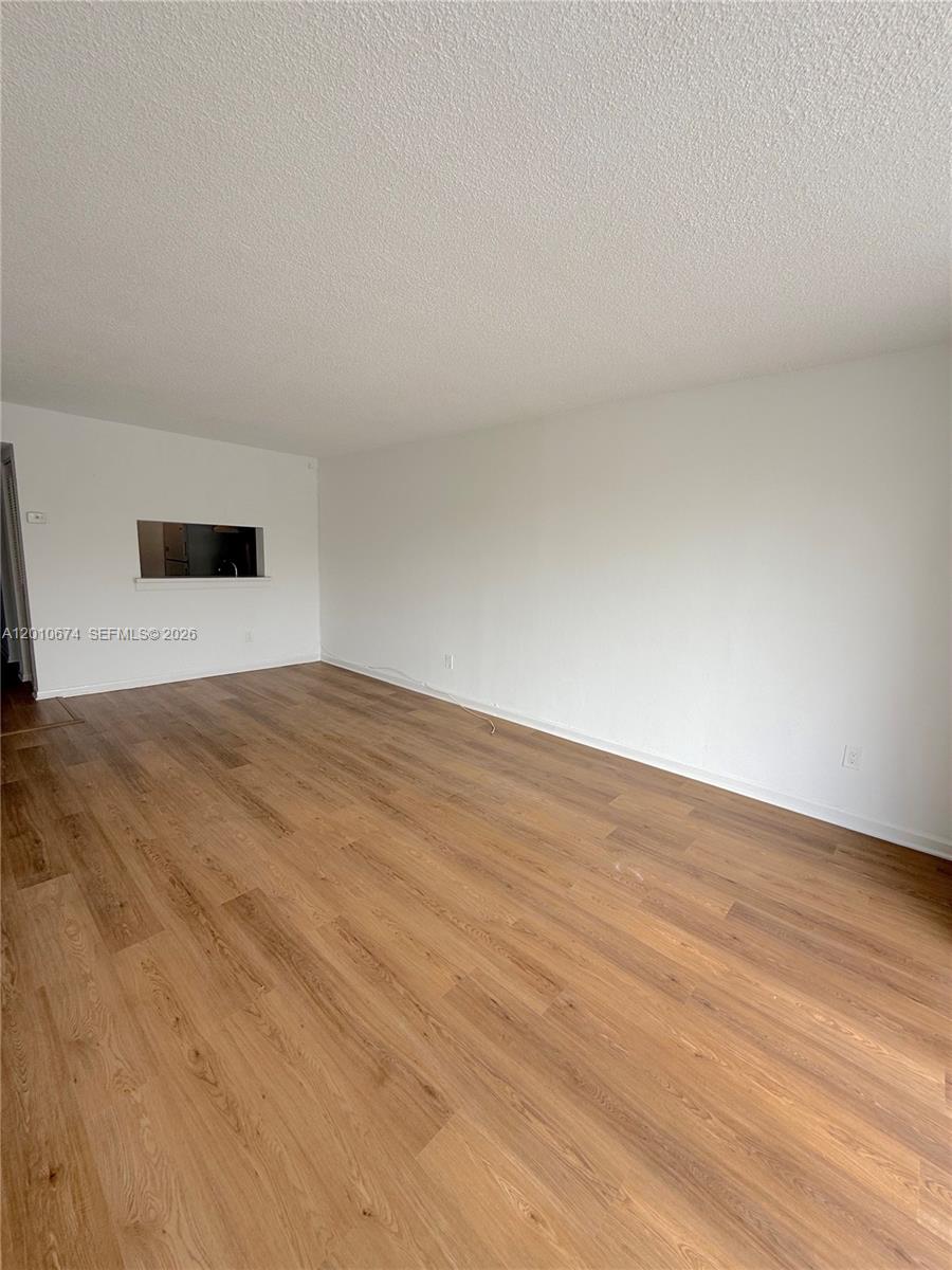 An empty living room features light wood-look flooring, white walls, and a kitchen pass-through window — 482 NW 165th St Rd # 307, Miami FL 33169, MIAMI, FL