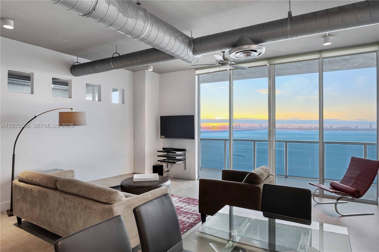 A modern living room features exposed ductwork, concrete ceilings, and floor-to-ceiling windows overlooking the ocean — 700 NE 25th St # 1902, Miami FL 33137, MIAMI, FL