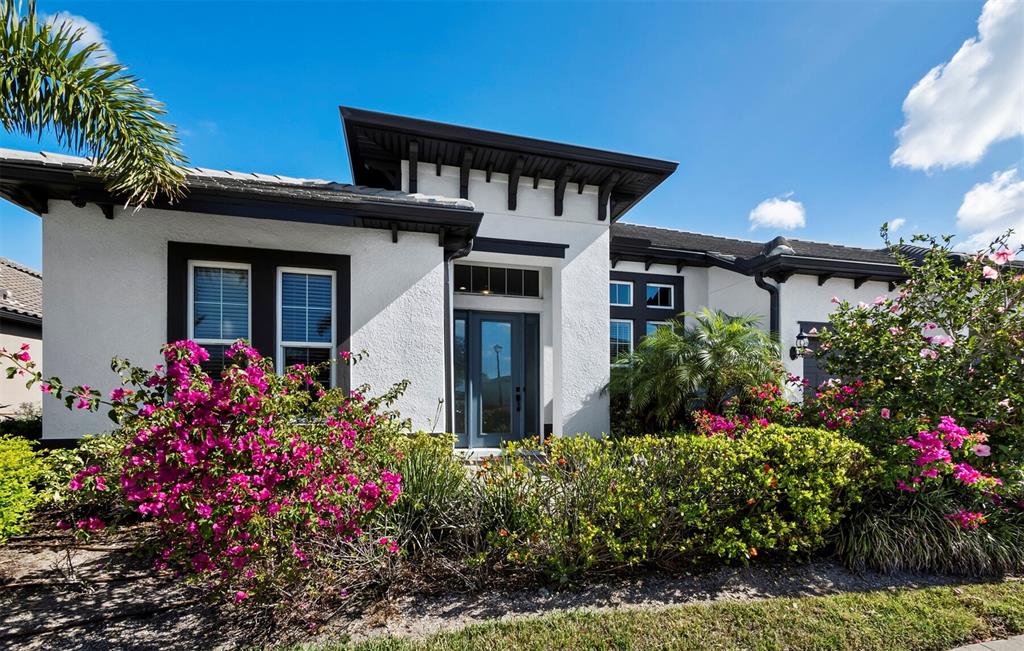 A single-story house with white stucco walls, dark trim, and vibrant bougainvillea bushes in front — 6129 LUNGO LAGO DRIVE, SARASOTA, FL