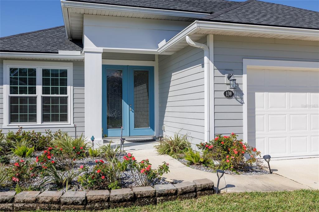 A gray house exterior features a blue double front door and a stone garden border — 139 WOOD STORK LANE, PALM COAST, FL