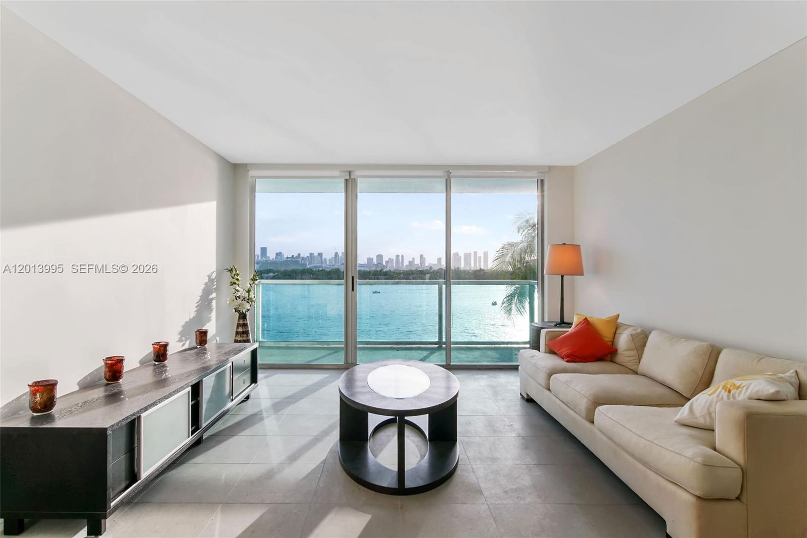 A living room with a beige sofa, circular coffee table, and floor-to-ceiling windows overlooking water — 650 West Ave # 805, Miami Beach FL 33139, MIAMI BEACH, FL