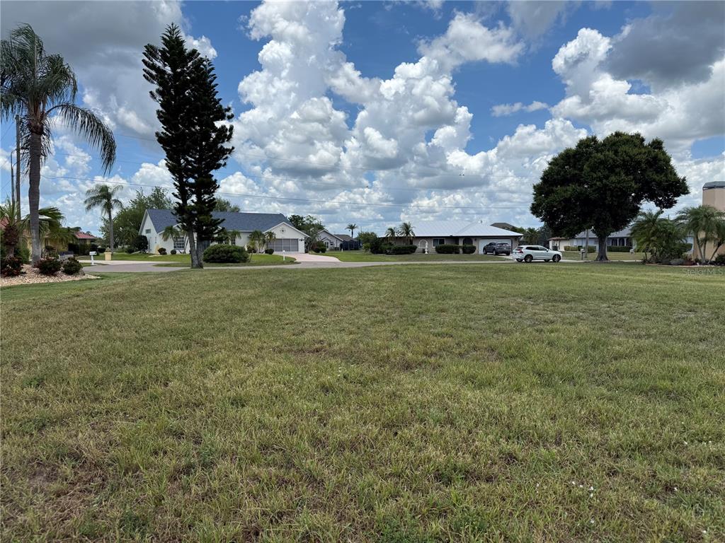 A large, grassy vacant lot is shown with residential houses and trees in the background — 25283 OJIBWAY COURT, PUNTA GORDA, FL