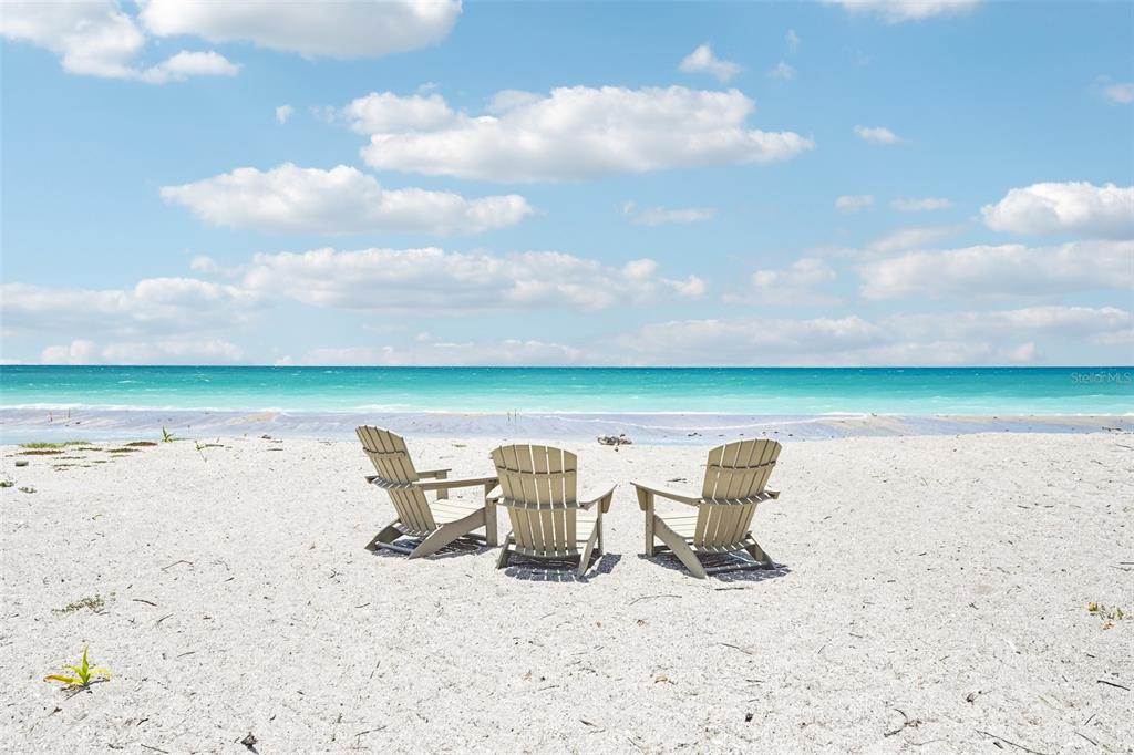 Three Adirondack chairs sit on a white sandy beach facing the turquoise ocean under clouds — 6570 MANASOTA KEY ROAD, ENGLEWOOD, FL