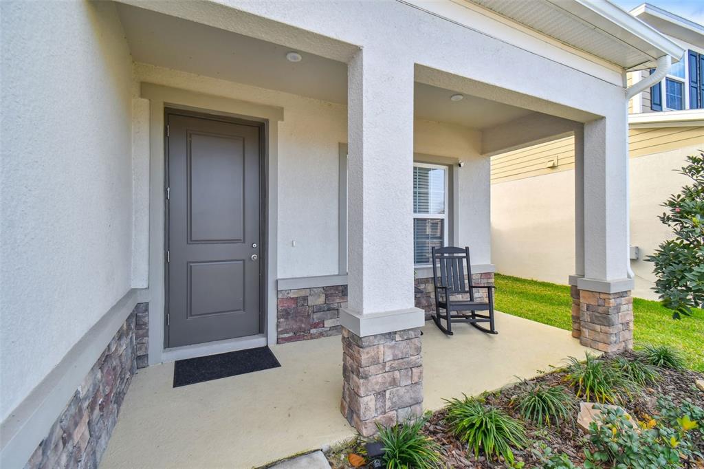 A covered front porch features a gray door, stone accents, and a black rocking chair — 8308 GREENWAY AVENUE, WESLEY CHAPEL, FL
