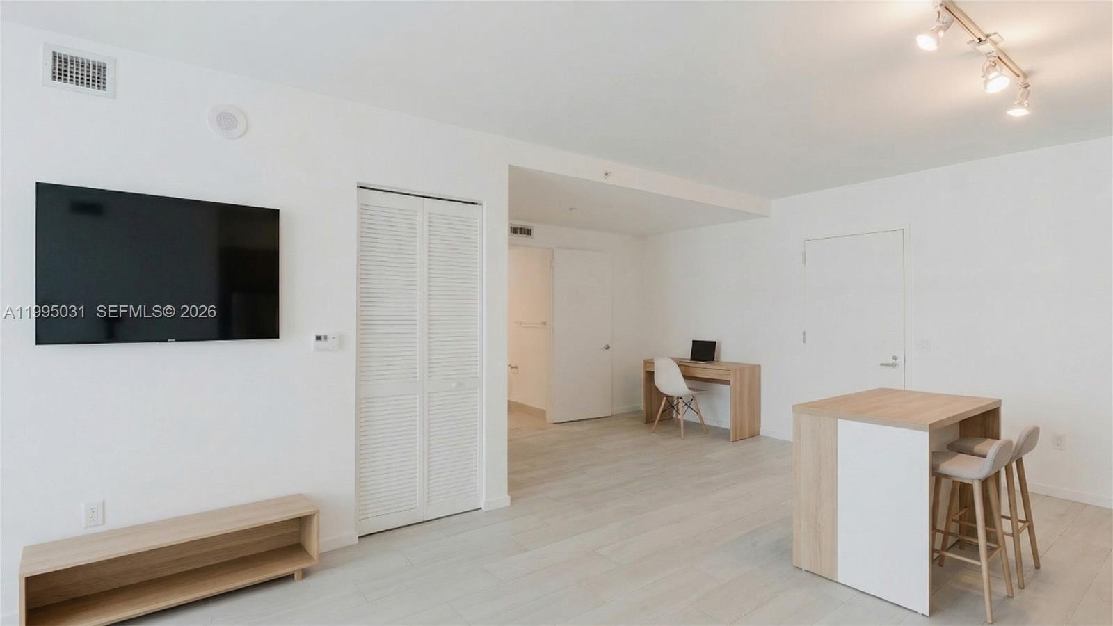 A bright, minimalist room features a wall-mounted television, a desk, and a small kitchen island — 31 SE 6th St # 1906, Miami FL 33131, MIAMI, FL