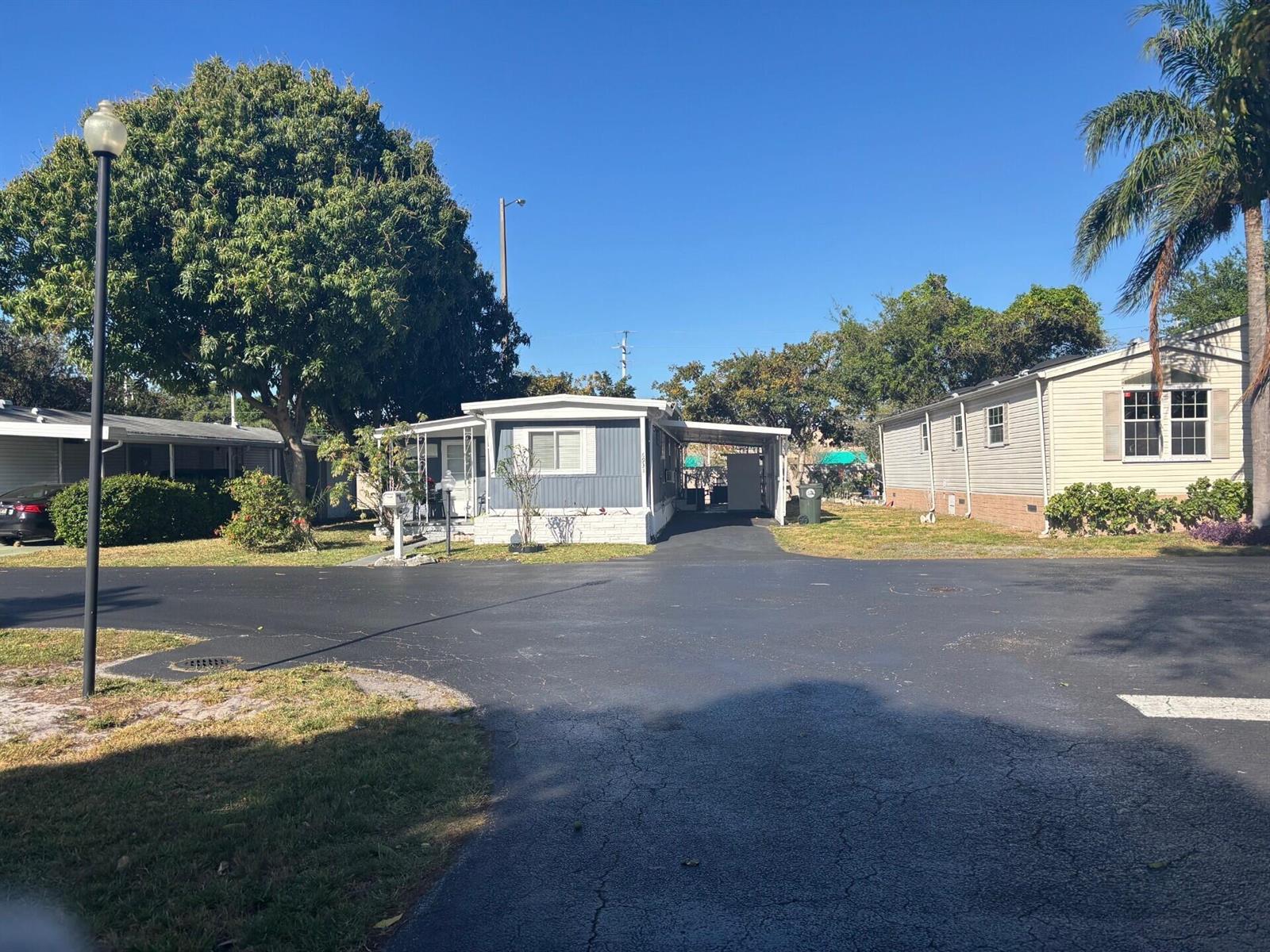 A blue mobile home with a carport sits on a paved lot with surrounding trees — 6931 NW 45th Terrace, Coconut Creek FL 33073, COCONUT CREEK, FL