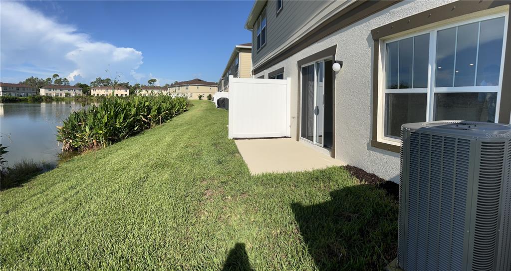 A view of a concrete patio, white privacy fence, and air conditioning unit beside a pond — 15031 CASPIAN TERN COURT UNIT 105, NORTH FORT MYERS, FL