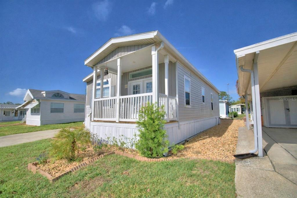 A light-colored mobile home with a front porch sits on a gravel and grass lot — 4925 CYPRESS GARDENS ROAD UNIT 100, WINTER HAVEN, FL