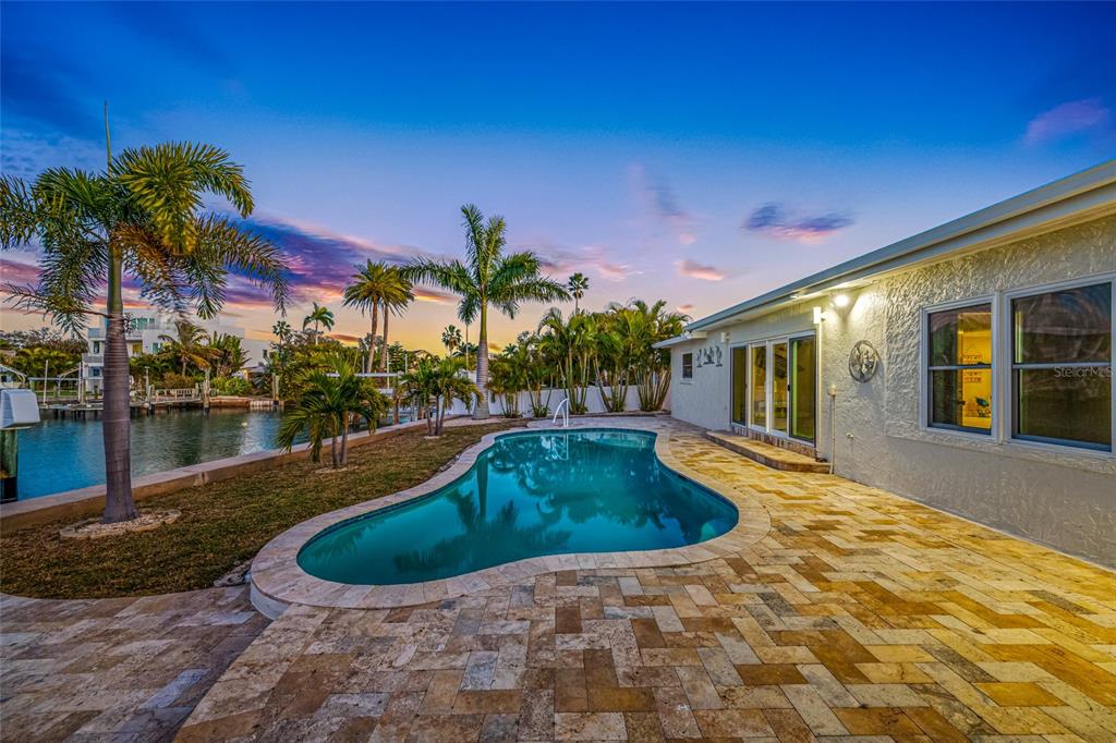 A backyard features a kidney-shaped swimming pool, stone patio, palm trees, and a waterfront view — 17161 2ND STREET E, NORTH REDINGTON BEACH, FL