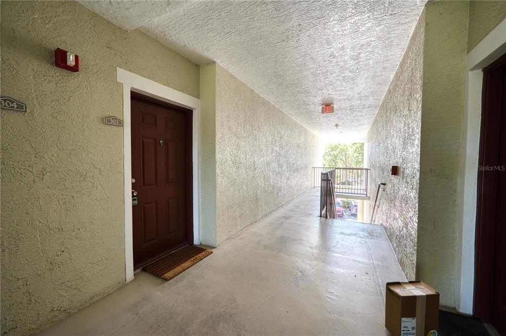 An outdoor apartment hallway features a brown door, concrete flooring, and a metal railing — 5551 BENTGRASS DRIVE UNIT 11-303, SARASOTA, FL