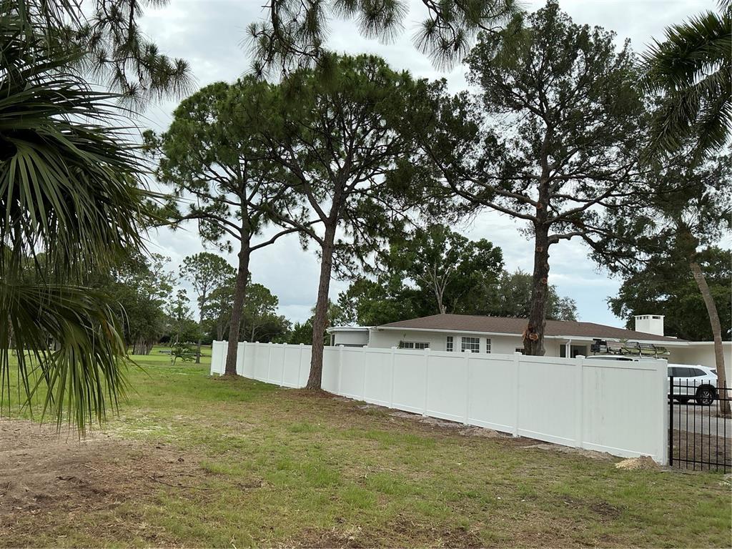 A white vinyl fence separates a grassy yard from a house with tall pine trees — 502 MAGELLAN DRIVE, SARASOTA, FL