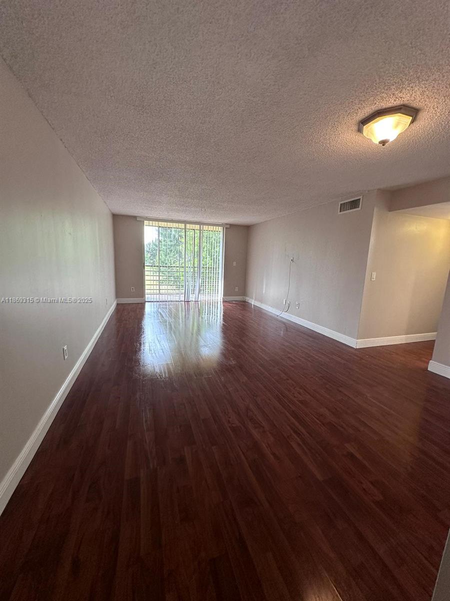 An empty living room features dark wood-look flooring, beige walls, and a sliding glass door — 3390 Foxcroft Rd # C212, Miramar FL 33025, MIRAMAR, FL