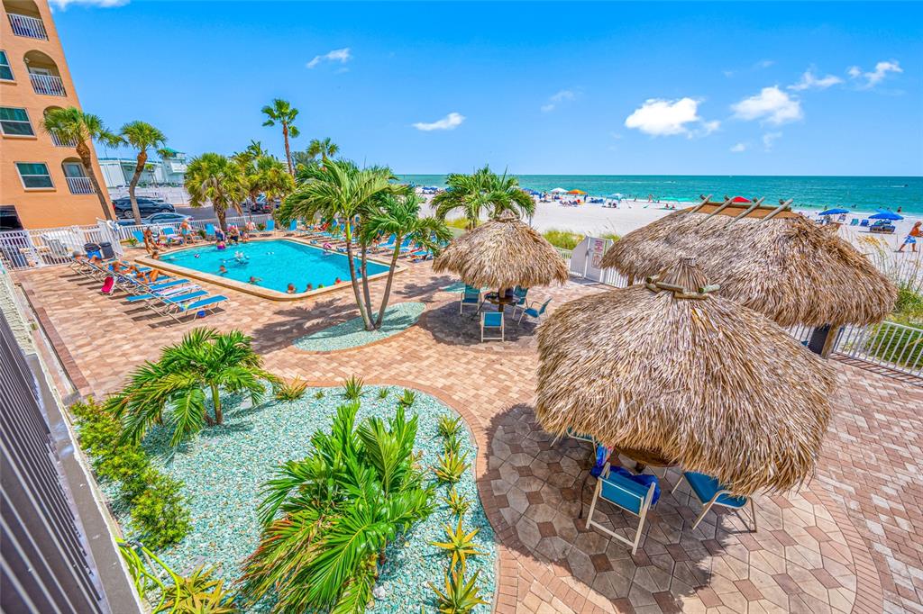 An elevated view of a resort pool area with tiki huts overlooking a sandy beach — 12924 GULF BOULEVARD UNIT 105, MADEIRA BEACH, FL