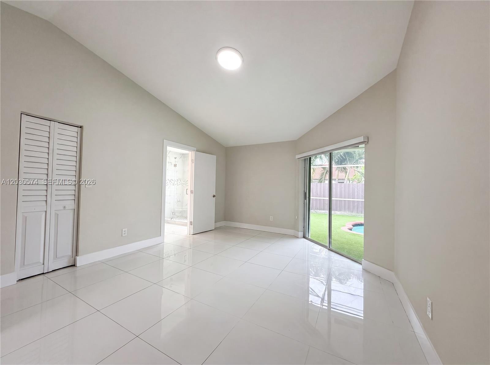 A bright, empty room features white tile floors, a vaulted ceiling, and sliding glass doors — 16571 SW 145th Ave, Miami FL 33177, MIAMI, FL
