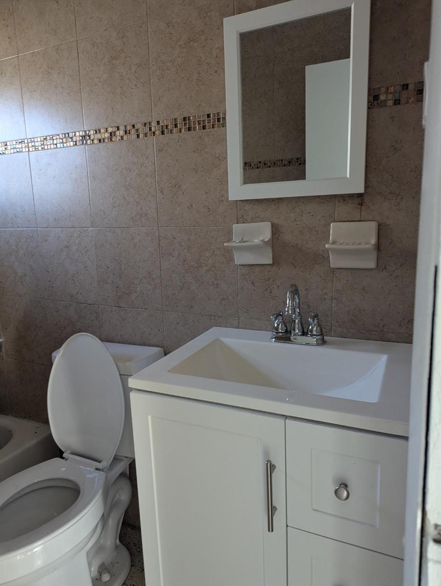 A bathroom featuring a white vanity, a wall-mounted mirror, and a toilet against tiled walls — 2736 Seneca Avenue, Fort Pierce FL 34945, FORT PIERCE, FL