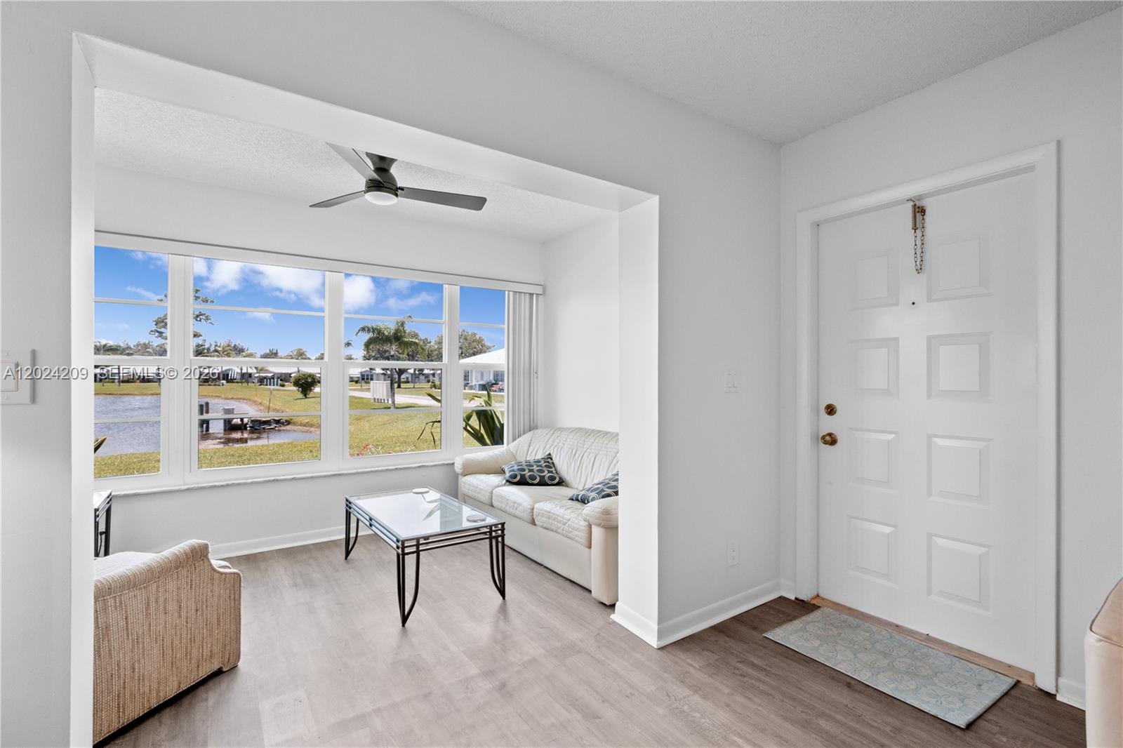 A living room features light wood-look flooring, a white sofa, and large windows overlooking water — 14604 Canalview Dr # B, Delray Beach FL 33484, DELRAY BEACH, FL