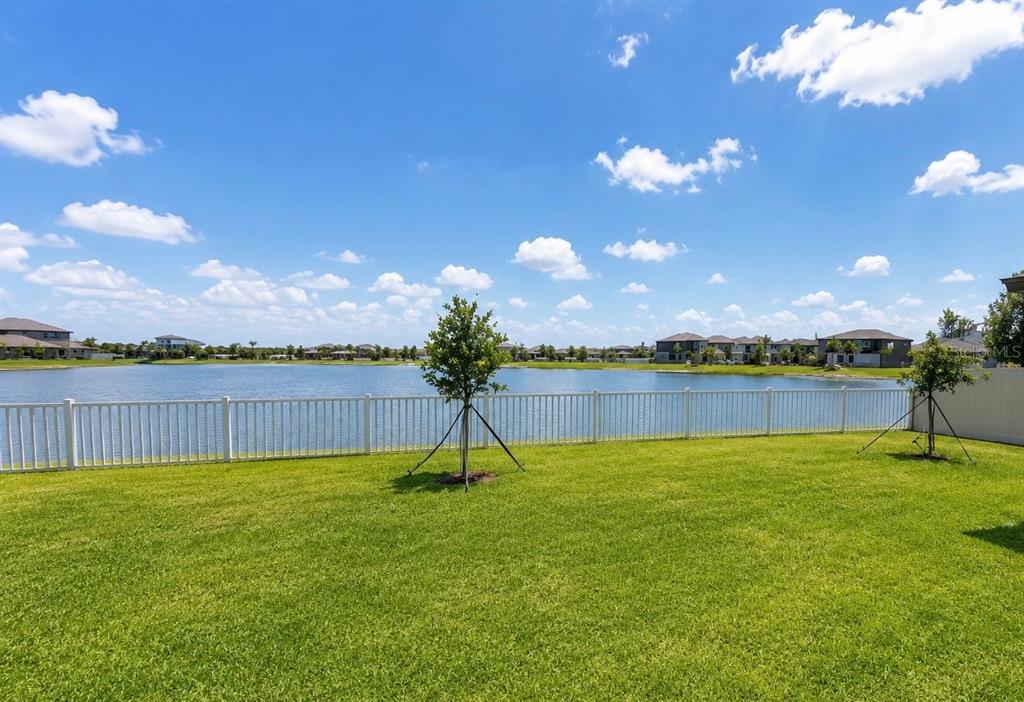 A grassy backyard features a white fence overlooking a lake under a blue sky — 12215 RADIANT GEM TRAIL, PARRISH, FL