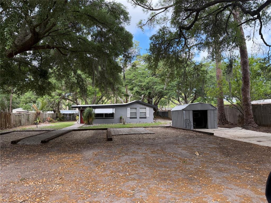 A single-story gray house with a metal shed sits on a large, tree-shaded dirt lot — 1257 LYNCHBURG ROAD, LAKE ALFRED, FL