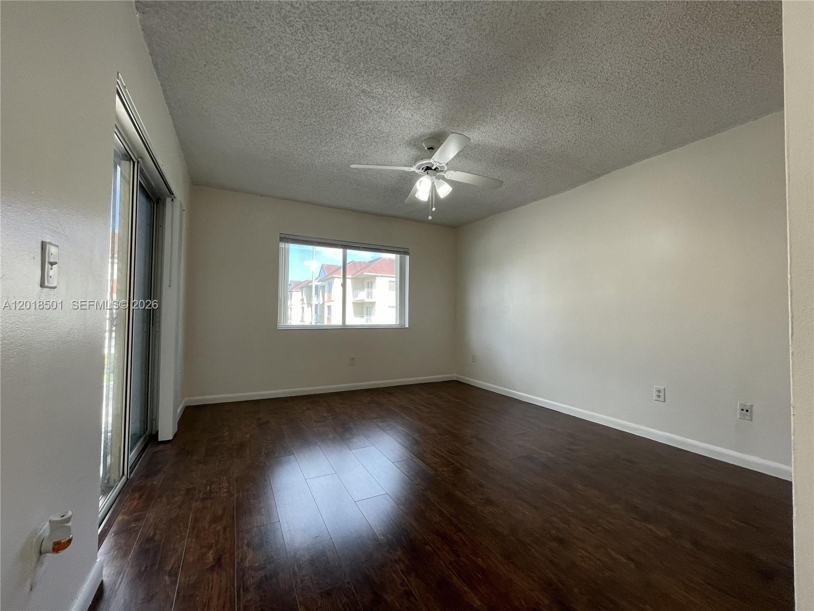 An empty room features dark wood-look flooring, white walls, a ceiling fan, and sliding glass doors — 18890 NW 57th Ave # 207, Hialeah FL 33015, HIALEAH, FL
