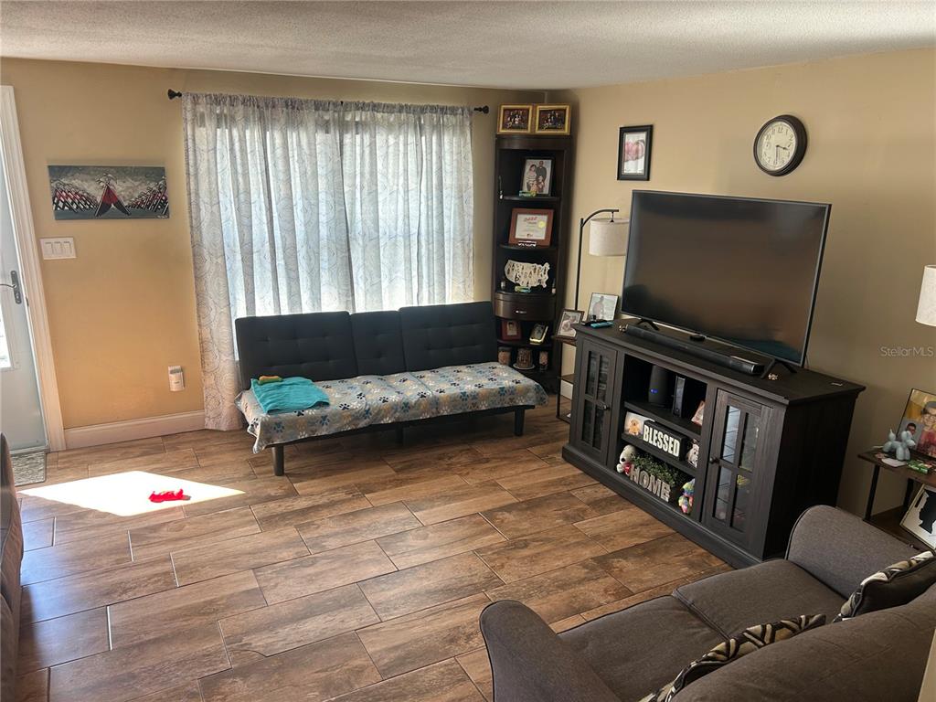 A living room features wood-look tile flooring, a black futon, and a television on a console — 10824 INGLEWOOD AVENUE, PORT RICHEY, FL