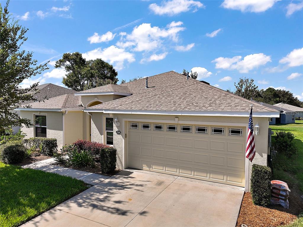 A single-story house with a beige stucco exterior, a two-car garage, and an American flag — 27102 CAMERONS RUN, LEESBURG, FL