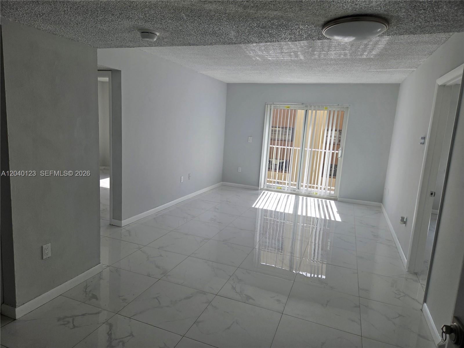 An empty room features light gray walls, white marble-patterned tile flooring, and a sliding glass door — MIAMI, FL