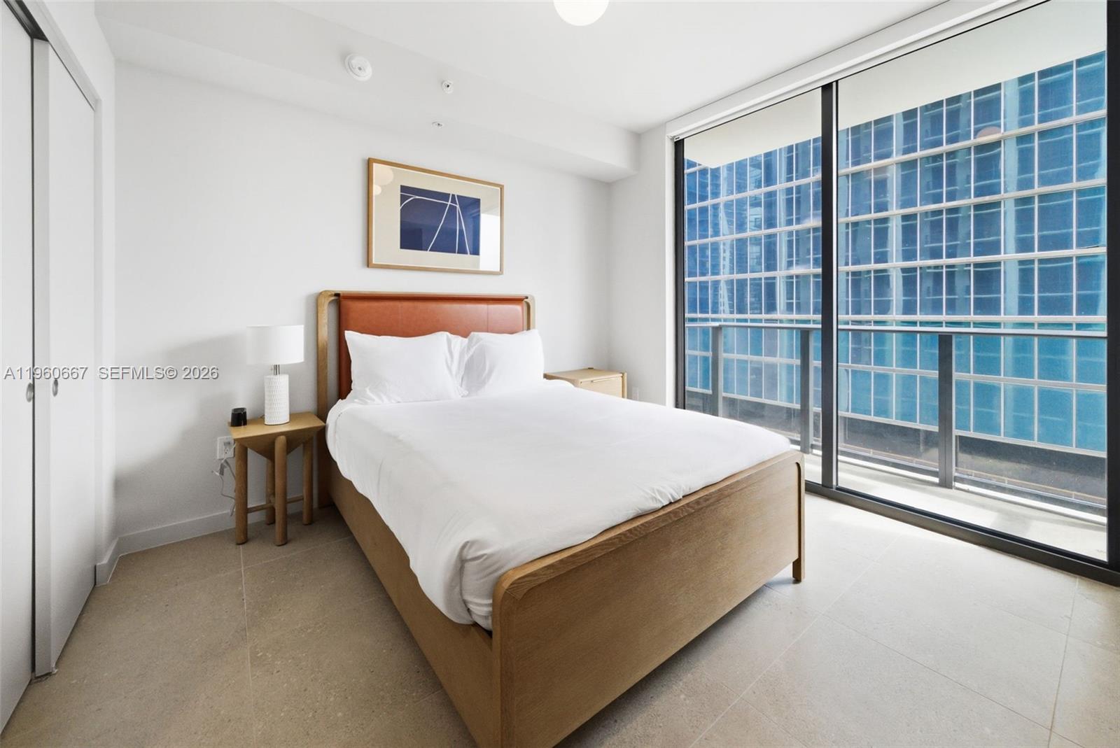 A bedroom features a wooden bed frame, white bedding, and floor-to-ceiling windows overlooking a building — 11 NE 6th St # 1802, Miami FL 33132, MIAMI, FL