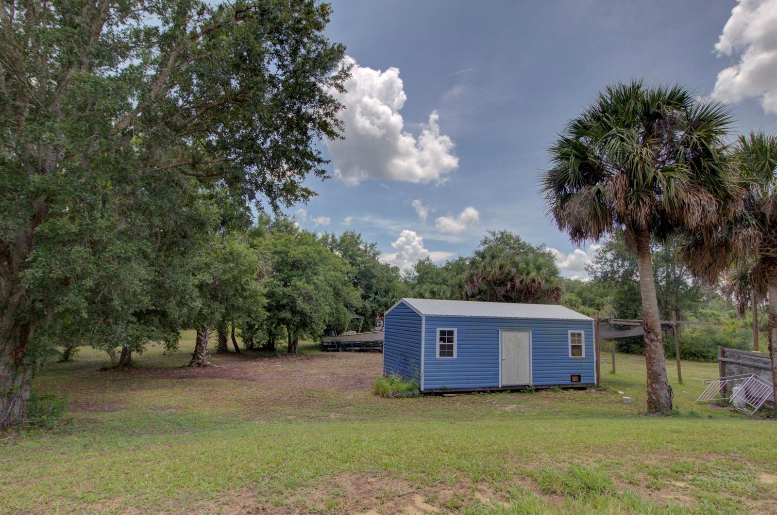 A blue storage shed sits in a grassy yard surrounded by trees and a palm tree — 19212 NW 280th Street, Okeechobee FL 34972, OKEECHOBEE, FL