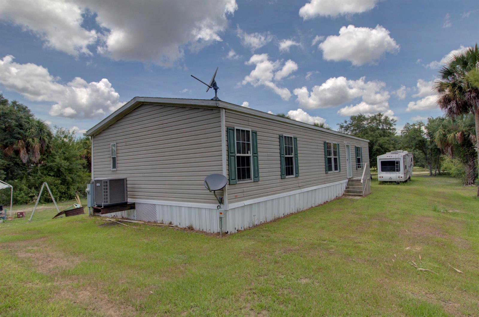 A beige mobile home with green shutters sits on a grassy lot under a cloudy sky — 19212 NW 280th Street, Okeechobee FL 34972, OKEECHOBEE, FL
