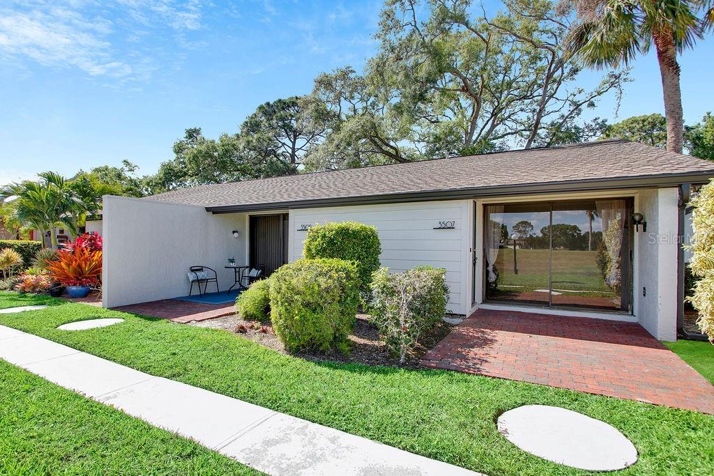 A single-story home exterior features a brick patio, sliding glass doors, and manicured landscaping — 3507 LONGMEADOW UNIT 4, SARASOTA, FL