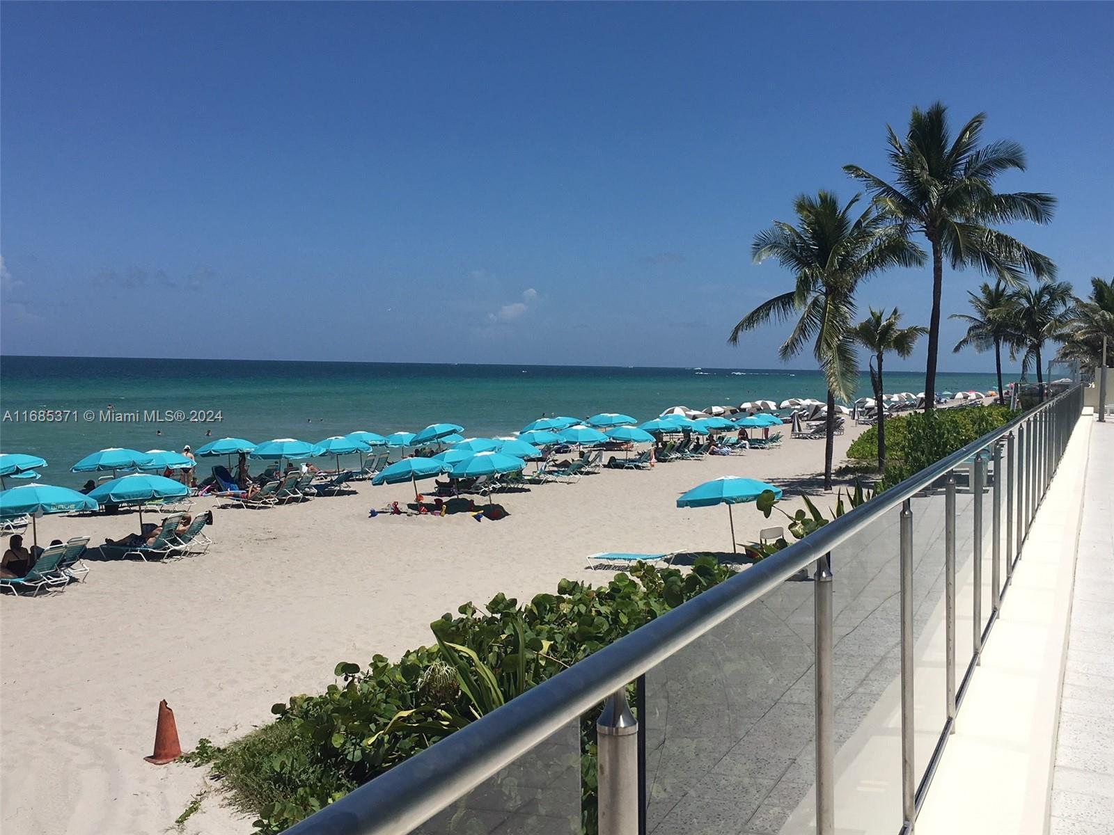 A view of a sandy beach with blue umbrellas and palm trees from a balcony — 2501 S OCEAN DR, Hollywood FL 33019, HOLLYWOOD, FL