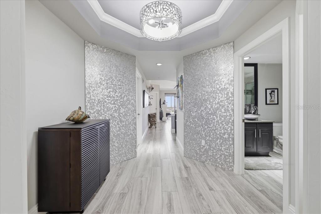A hallway features light wood-look flooring, two silver mosaic accent walls, and a decorative chandelier — 14300 RIVA DEL LAGO DRIVE UNIT 1802, FORT MYERS, FL