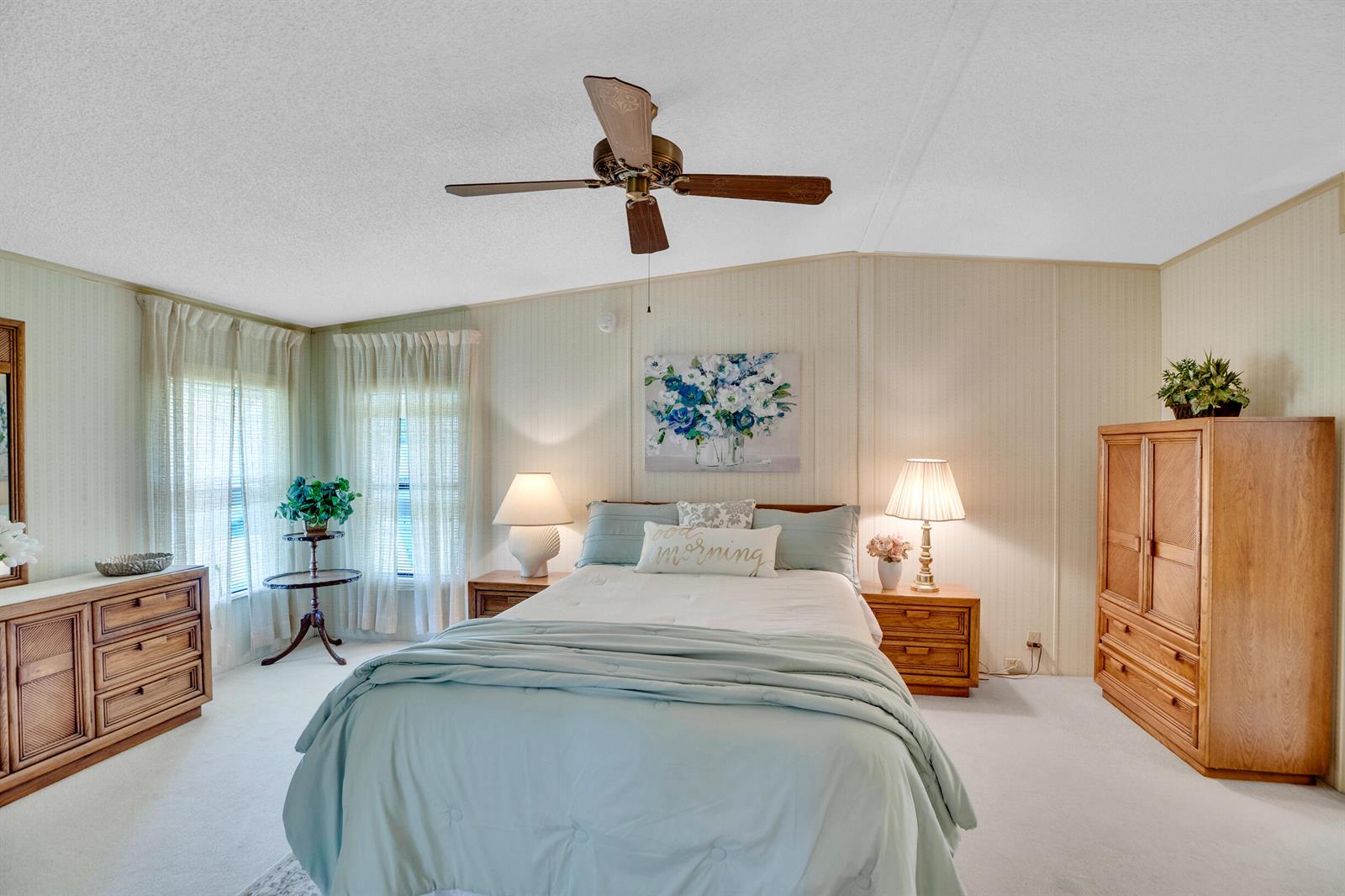A bedroom features a queen bed, light wood furniture, a ceiling fan, and light-colored walls — FL