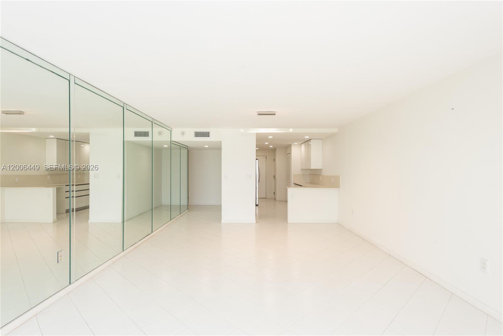 An empty room features white tiled floors, a mirrored wall, and an adjacent kitchen area — 600 Grapetree Dr, Key Biscayne FL 33149, KEY BISCAYNE, FL