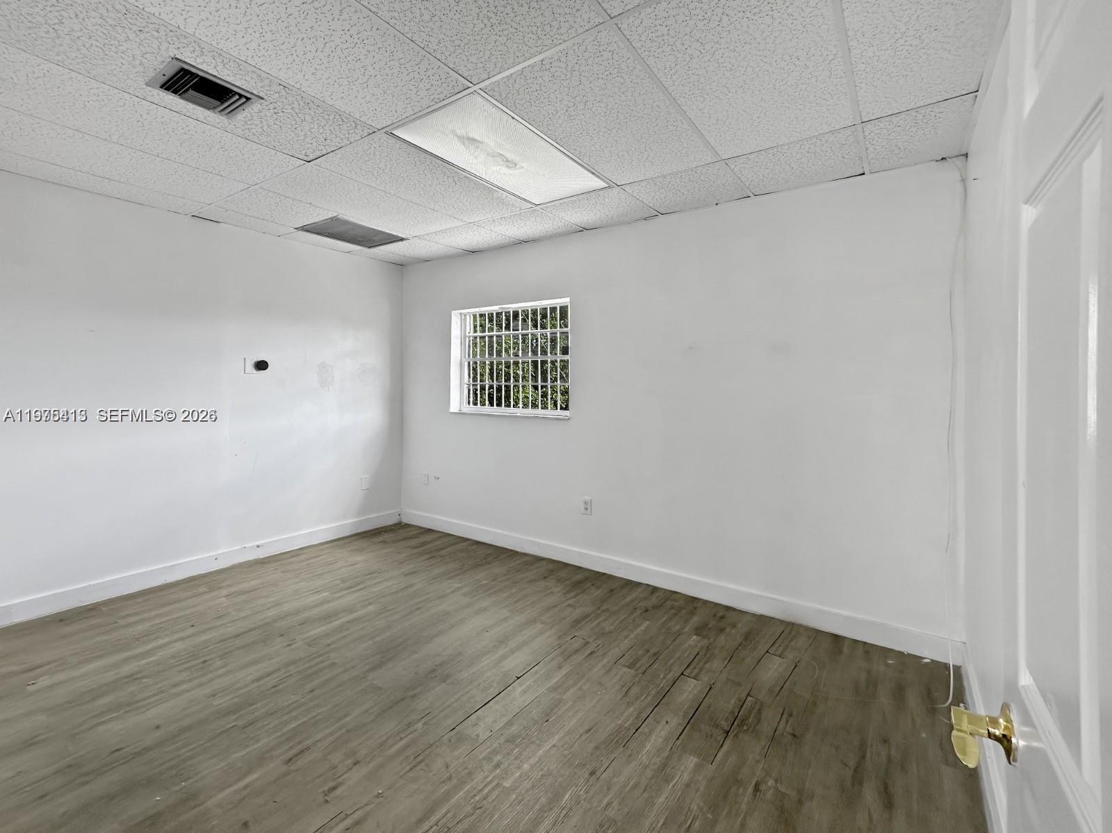 An empty room features white walls, wood-look flooring, a drop ceiling, and a barred window — 497 NW 27th Ave # 497, Miami FL 33125, MIAMI, FL