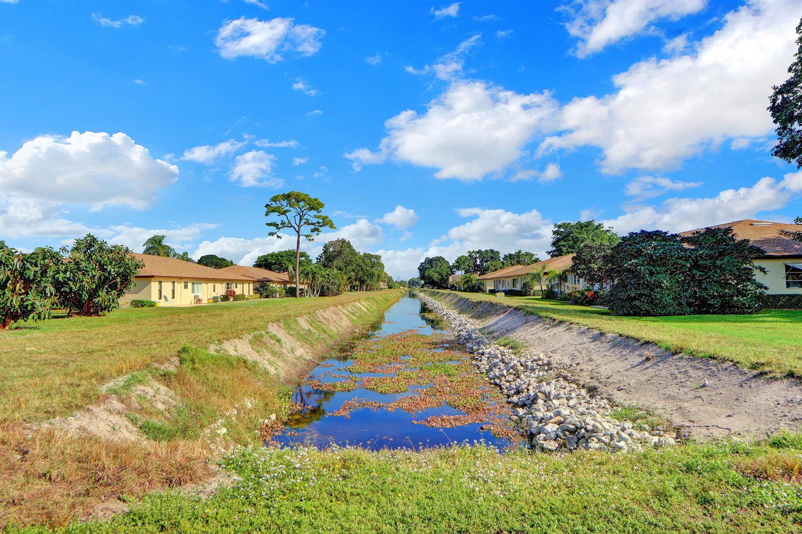 A canal runs between two rows of single-story residential buildings under a blue sky — 14050 Nesting Way # B, Delray Beach FL 33484, DELRAY BEACH, FL