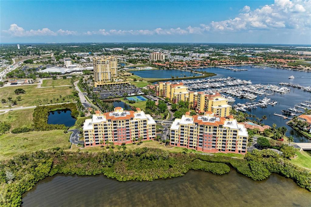 An aerial view shows waterfront apartment buildings, a marina, and surrounding landscape under a blue sky — 611 RIVIERA DUNES WAY UNIT 403, PALMETTO, FL