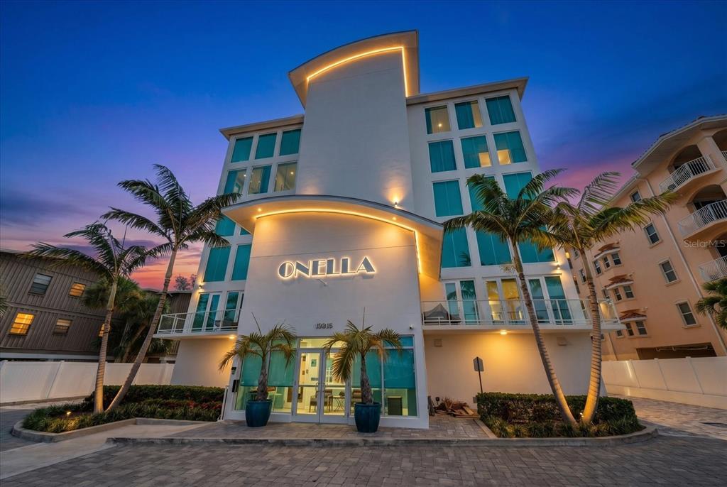 Exterior view of a modern multi-story building with palm trees and illuminated signage at dusk — 19816 GULF BOULEVARD UNIT 501, INDIAN SHORES, FL