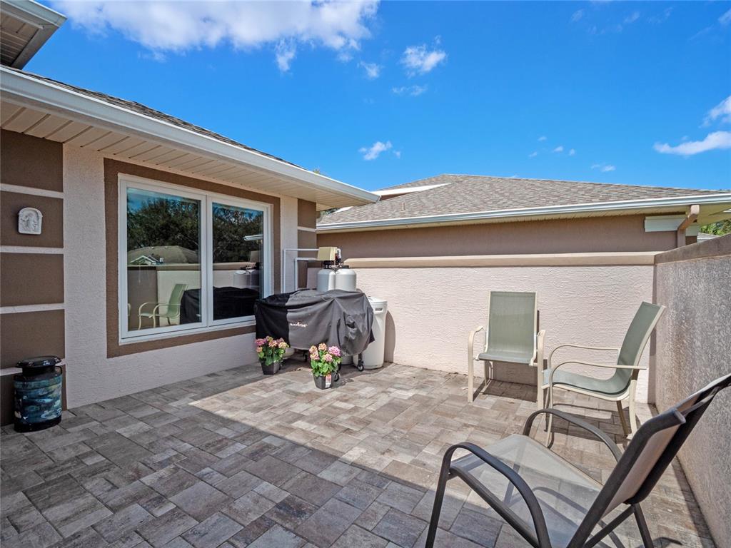 A paved patio features a covered grill, potted flowers, and outdoor chairs against a wall — 21849 TARTAN STREET, LEESBURG, FL