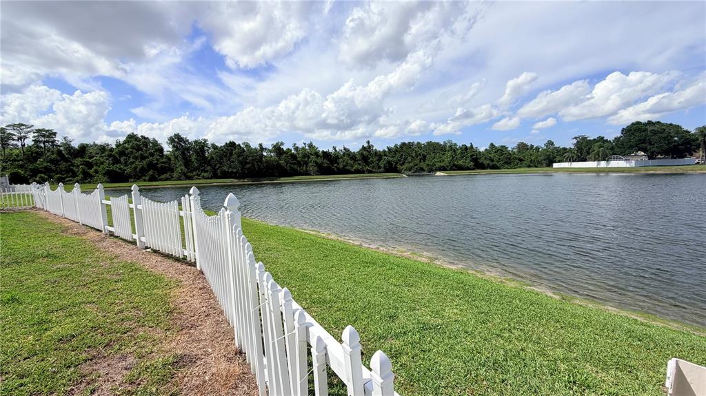 A white picket fence borders a grassy lawn next to a lake under a cloudy sky — 3113 FAIRFIELD DRIVE, KISSIMMEE, FL