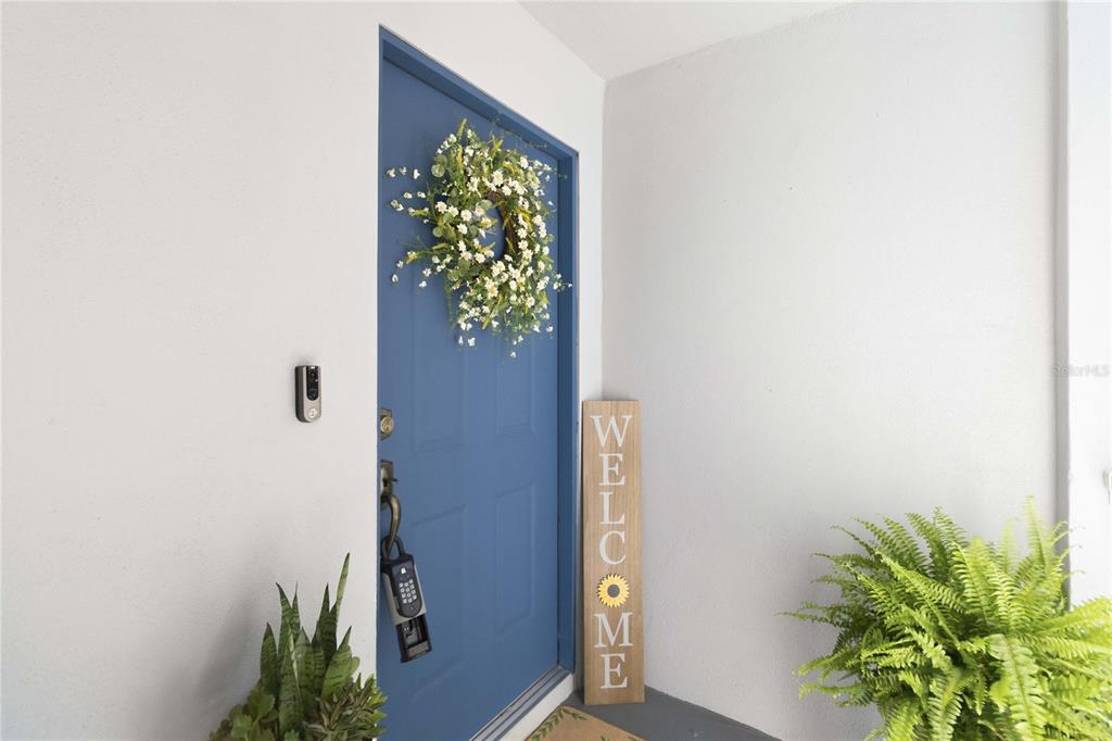 A blue front door with a wreath and welcome sign sits between two potted plants — 6206 BAYHILL LANE UNIT 6206, LONGWOOD, FL